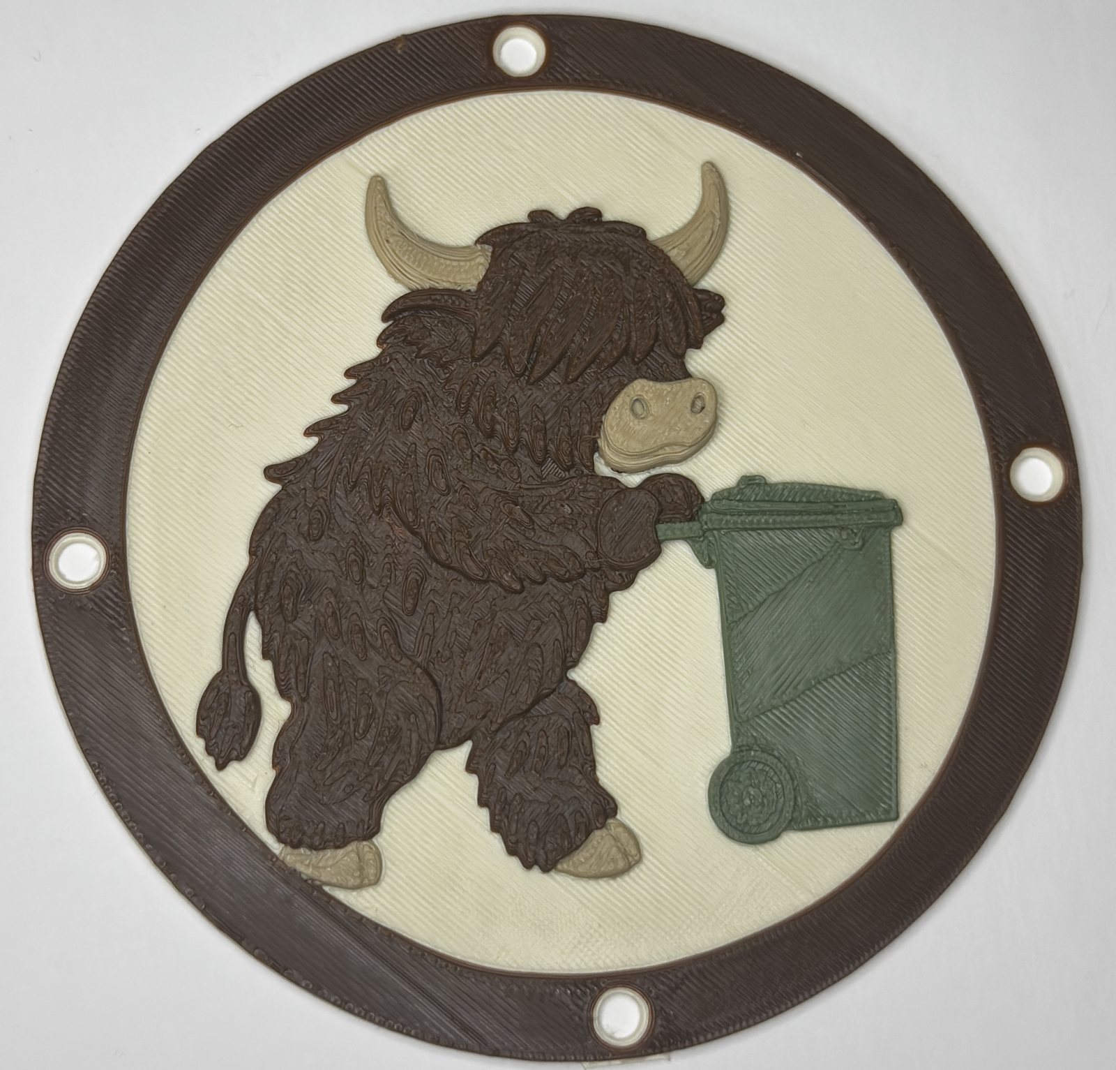 3D printed circular bin wheel trim with a multi-colour Highland cow design, showing a shaggy brown Highland cow pushing a green wheelie bin on a cream background with a brown border and screw holes